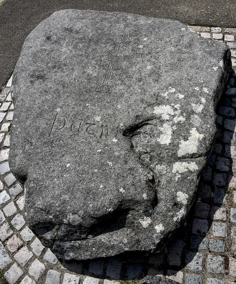 A large rough granite boulder set in cobblestones, carved with a small cross and the name "Patric"