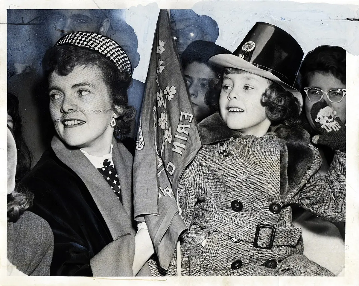 Black-and-white photograph of a smiling mother and her six-year-old daughter in a top hat holding Erin Go Bragh banners as they watch the parade on Fifth Avenue
