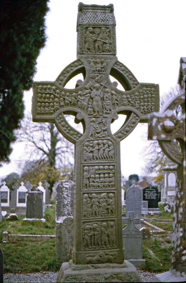 A tall carved stone high cross with a ringed head, every panel of its shaft and arms filled with carved scenes from Scripture, standing in an Irish graveyard