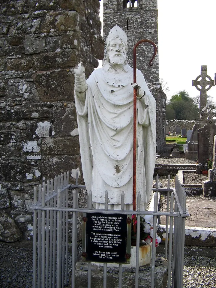 White statue of St. Patrick in bishop's vestments and miter, raising his right hand and holding a crozier, inside a low iron railing in front of a ruined stone tower