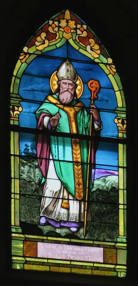 Tall arched stained-glass window of St. Patrick in a green cope and miter holding a crozier, standing in a green landscape under a decorated Gothic canopy