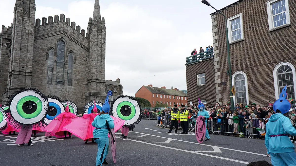 Performers in bright pink costumes carrying giant round eye puppets march past crowds behind barriers in a Dublin street, with a church spire behind