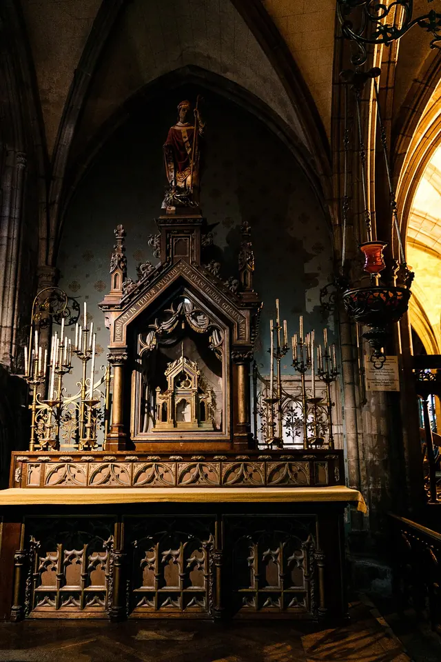 A carved dark wooden Gothic altar in a side chapel, with a gilded reliquary shrine in a canopied niche and a statue above, lit by candles