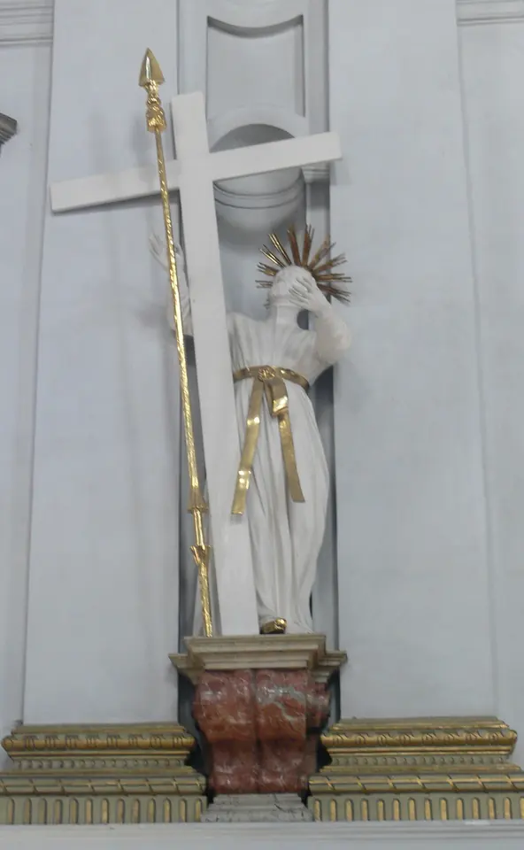White statue of a young man with a gold halo, wearing a long white robe with a gold sash, leaning against a tall cross with a gilded spear beside him