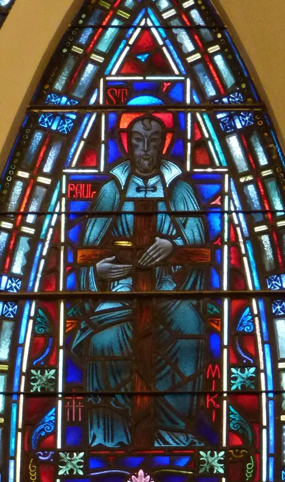 Pointed stained-glass window of a Japanese saint in a dark robe with a halo, holding a cross before him, with the words Paul and Miki in red letters