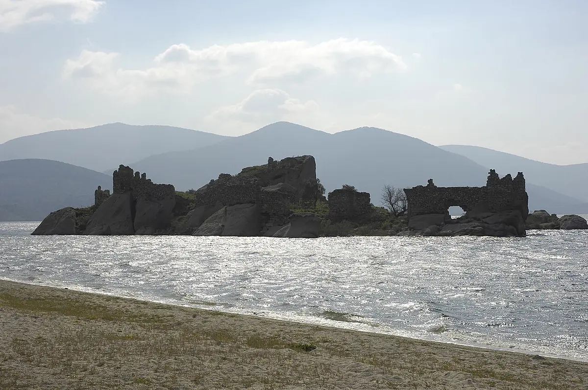 Ruined stone walls of a medieval monastery covering a small rocky island in a lake, with blue mountains beyond