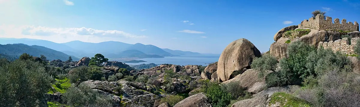 Wide view across rounded granite boulders and green scrub to the ruined walls of a monastery on a rocky ridge, with Lake Bafa and distant hills beyond