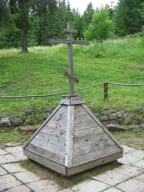 A wooden Orthodox cross on a small pyramid-shaped wooden well cover, with a grassy slope and fir trees behind