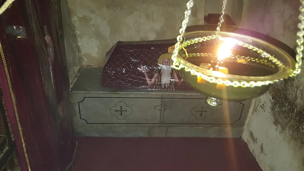 A low stone tomb draped with a red embroidered cloth in a small rock-cut chamber, lit by a hanging oil lamp