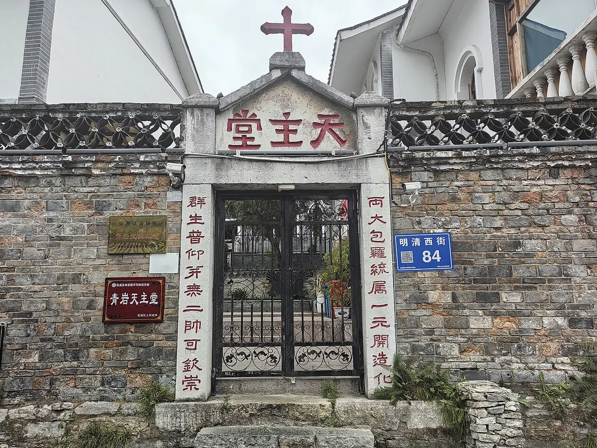 Gray brick wall with an old stone gateway topped by a red cross and red Chinese characters, iron gates, and white church towers behind