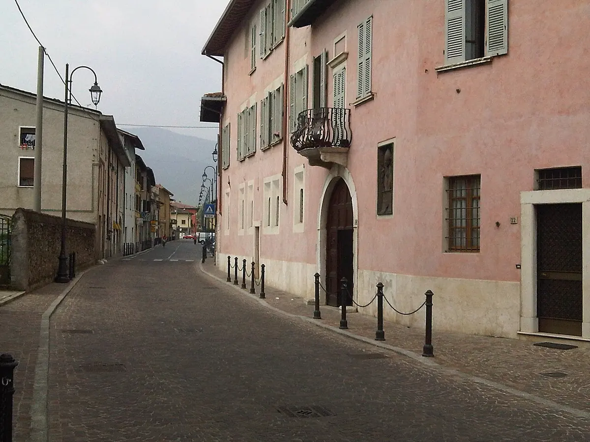 A pink-plastered three-story house with green shutters and a balcony on a quiet village street in Concesio