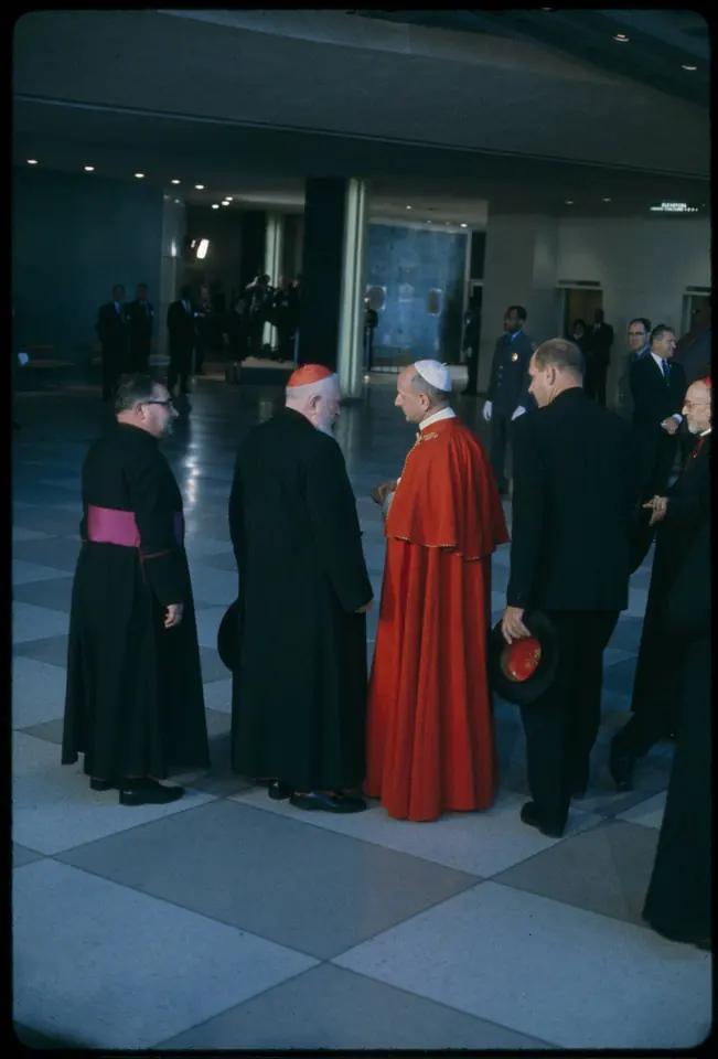 Color photograph of the pope in a red cape walking among clergy in black cassocks and a cardinal in red in a modern hall at the United Nations