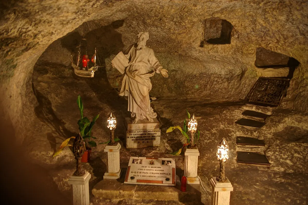 A rock-cut grotto with a white marble statue of St. Paul on a pedestal, lit by lamps and candles, with flowers at its base