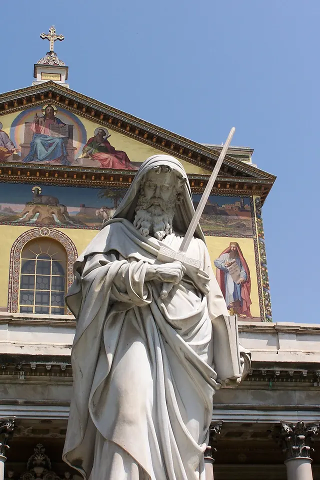 White marble statue of a bearded St. Paul holding a long upright sword, in front of the basilica's mosaic-covered facade