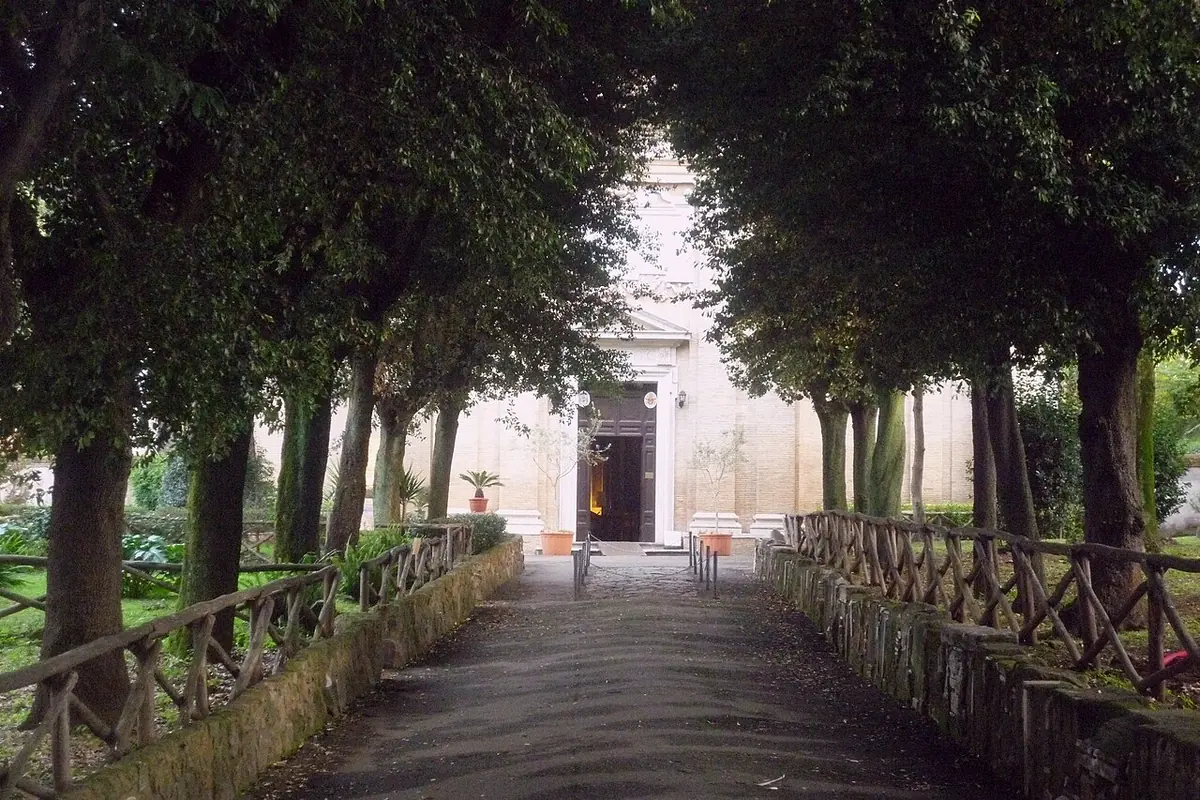 A tree-lined path leading to the pale stone facade of a small church in the grounds of the Tre Fontane abbey