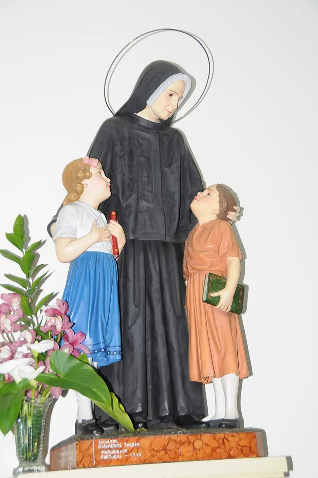 Painted statue of a nun in a black habit and white bonnet with a halo, standing between two small girls holding a book and a pencil