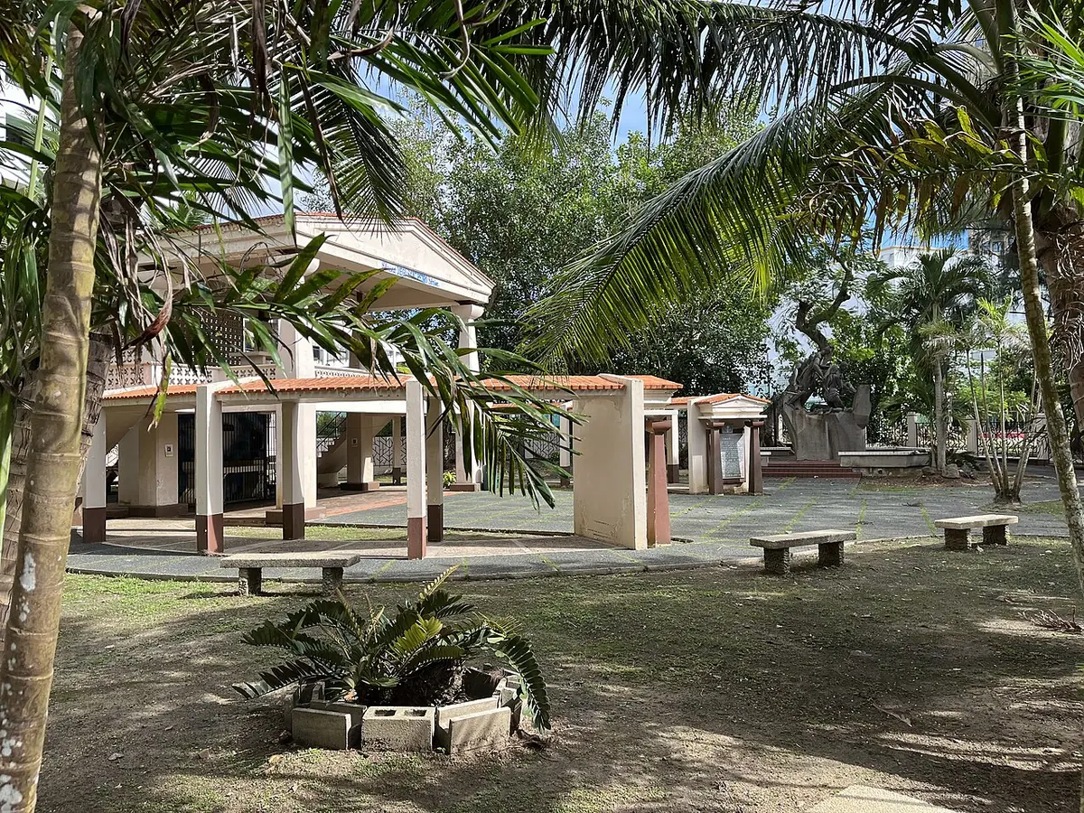 A small open-sided memorial pavilion with a tiled roof in a shady garden of palms and cycads at the site of the martyrdom in Tumon