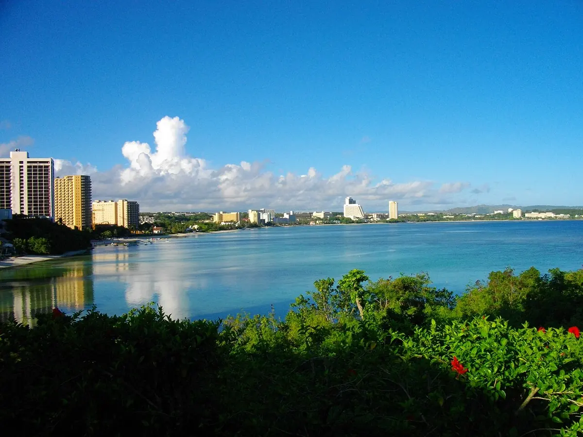 The calm turquoise water of Tumon Bay on Guam, curving toward a line of hotels under a bright blue sky with white clouds