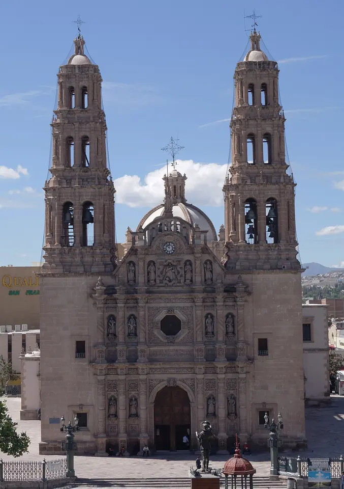 The carved stone front of a Baroque cathedral with two tall bell towers and a dome, above a wide plaza