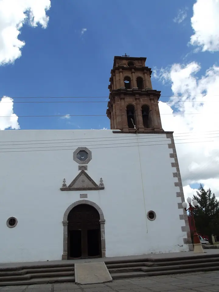 A white colonial church with a plain arched doorway, a small round window and a brick bell tower against a blue sky with white clouds