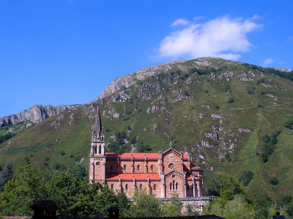 A pink stone neo-Romanesque basilica with twin spires set against a steep green mountainside under a blue sky