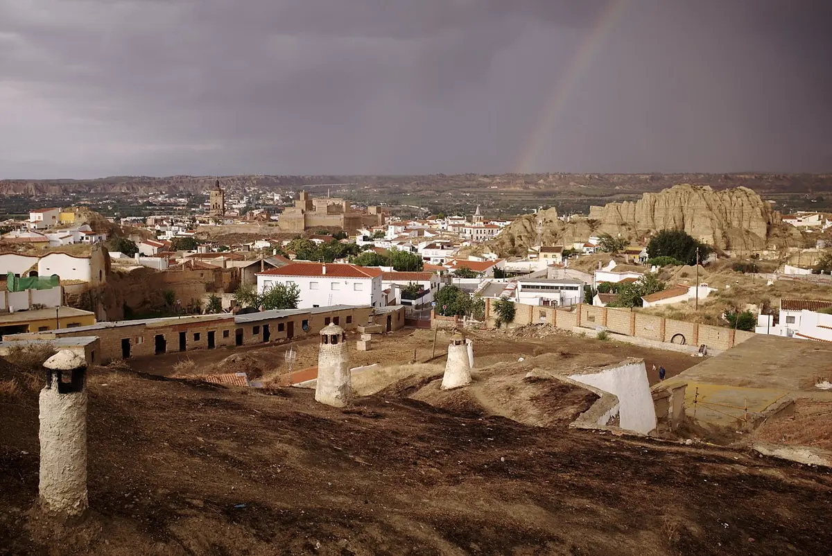 White cave chimneys rising from earthen roofs in the hills of Guadix, with the town and a rainbow under a stormy sky behind