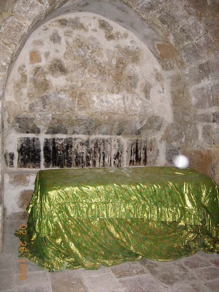 A stone tomb covered with a shiny green cloth in a low vaulted chamber of rough limestone blocks