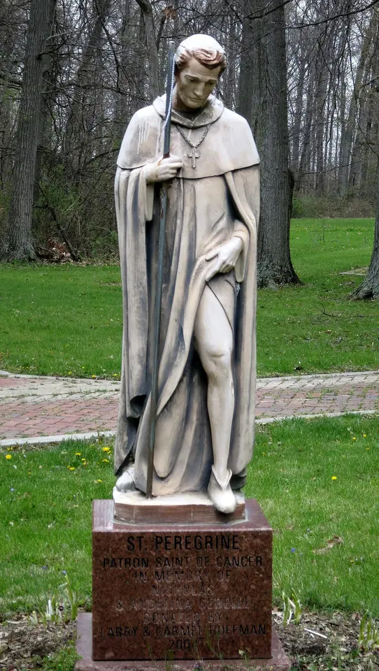 White stone statue of St. Peregrine in a hooded habit, holding a crucifix and lifting his robe to show his leg, on a red granite base in a wooded park