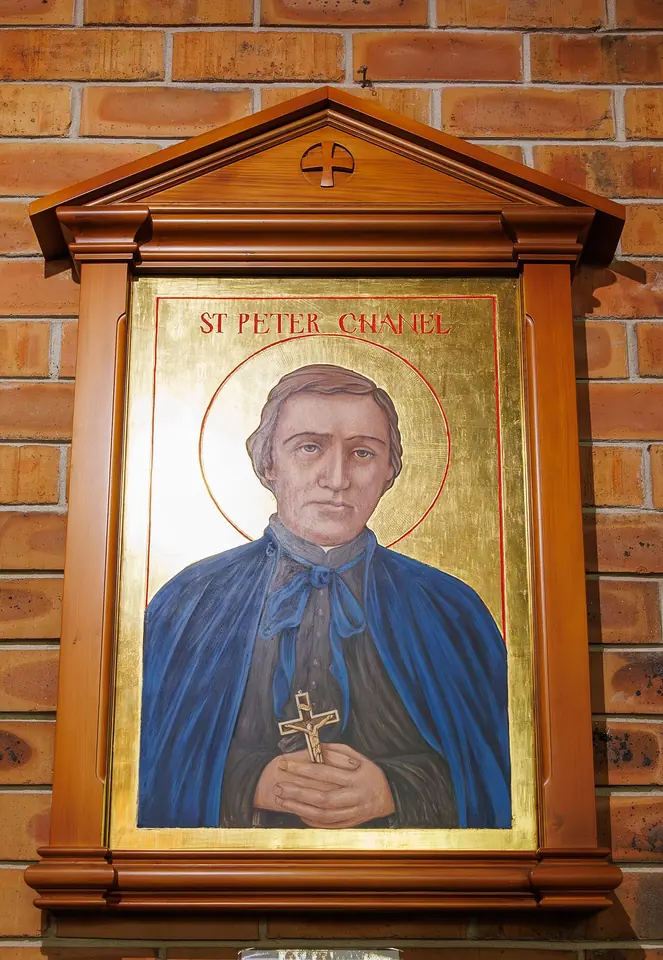 Gold-ground icon of St. Peter Chanel in a blue cloak over a black cassock, holding a small crucifix, in a carved wooden frame on a brick wall