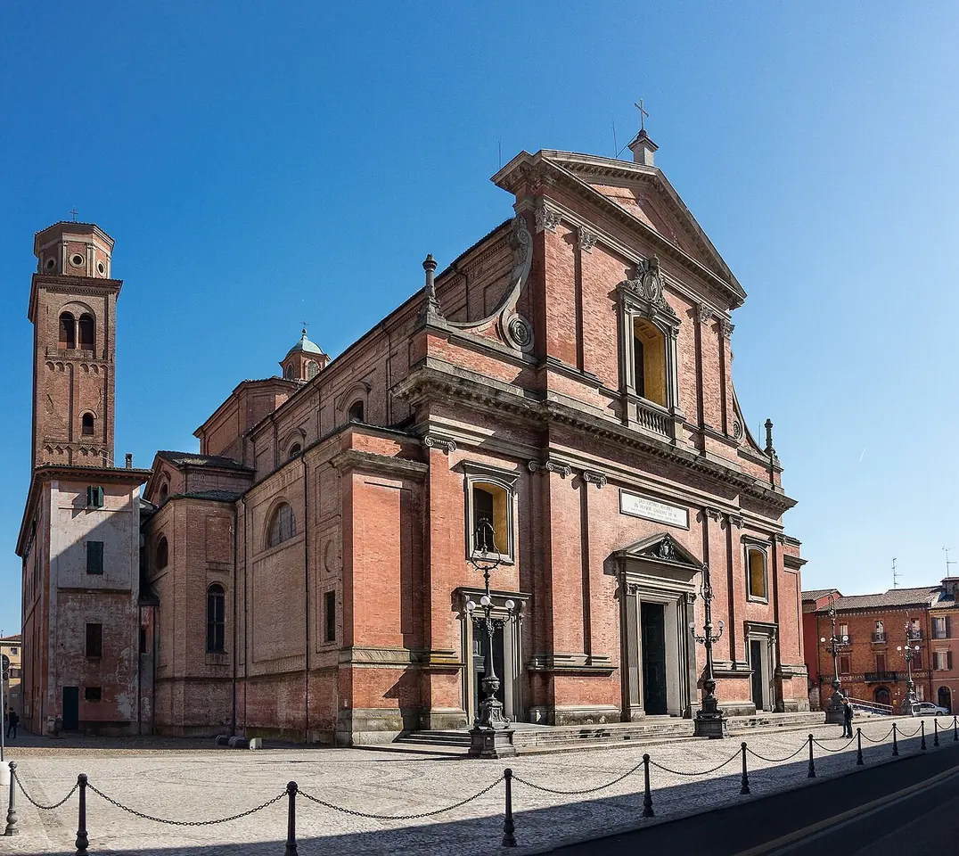 Red-brick facade of Imola's cathedral with classical white stone doorways and a tall brick bell tower on the left, under a clear blue sky