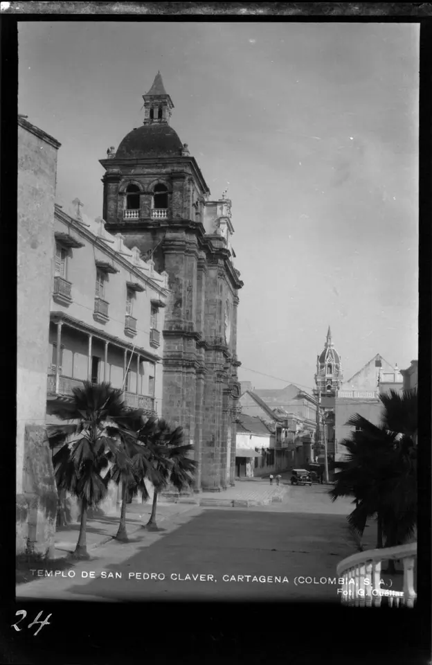 Fotografía en blanco y negro de la torre de piedra y la fachada de la iglesia de San Pedro Claver al final de una calle con palmeras y casas de balcones