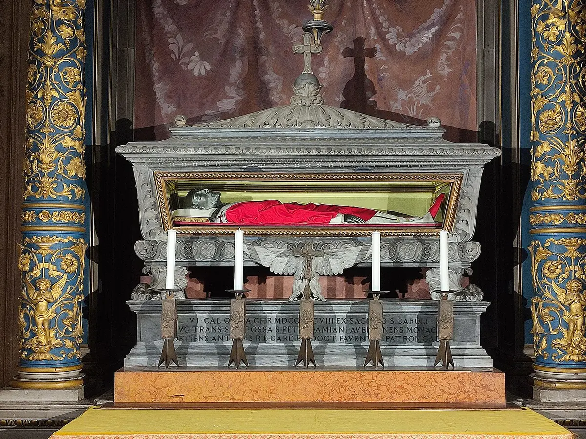 Gray stone shrine with a glass front holding the vested remains of St. Peter Damian in red, with six candles on the altar below and gilded columns on either side