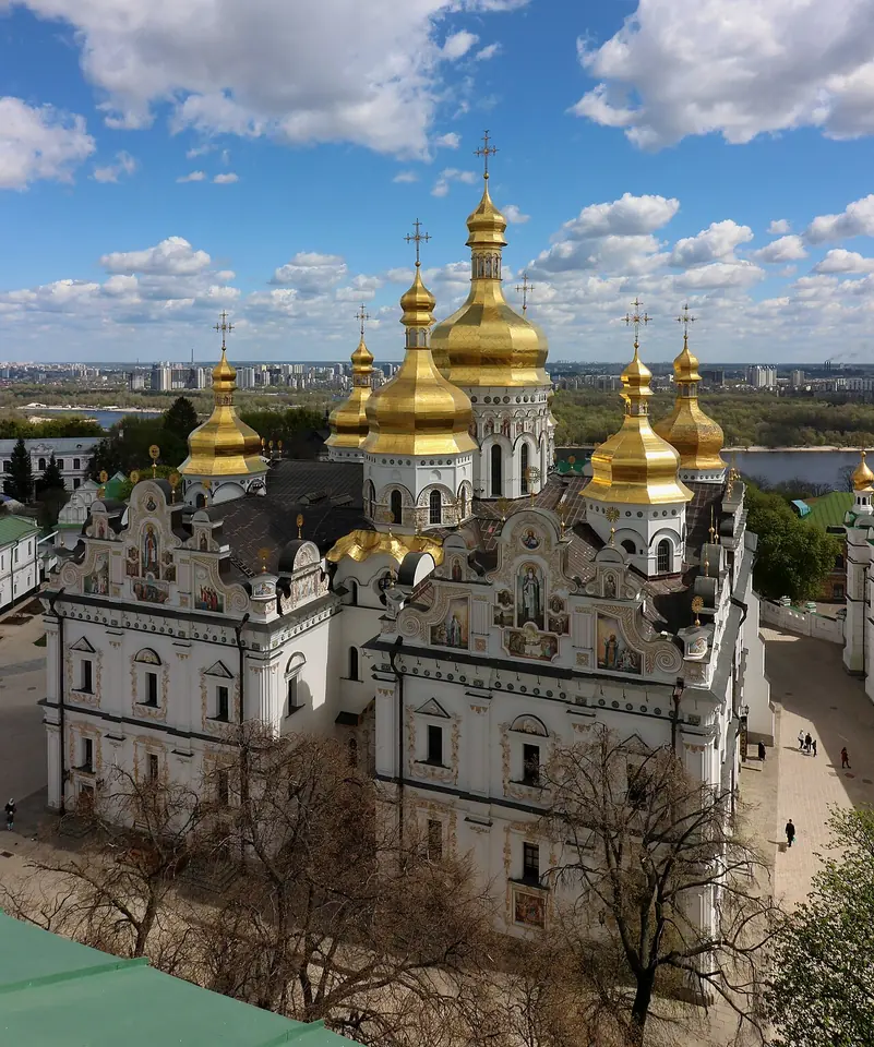 White Baroque cathedral with many golden domes and an ornate gabled façade, seen from above, with the Dnieper River in the distance