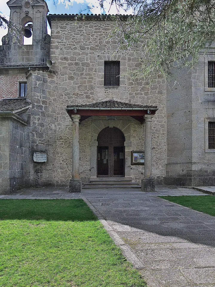 A simple granite church front with a small columned porch and a little bell gable, beside a lawn and a stone path