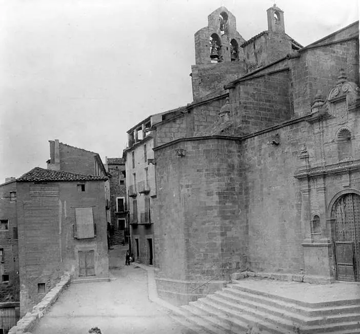 An old black-and-white photograph of a narrow village street climbing to a large stone church with a bell gable