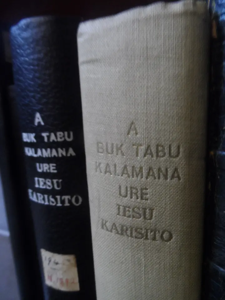 Spines of two old bound books on a library shelf, stamped in gold "A Buk Tabu Kalamana ure Iesu Karisito"