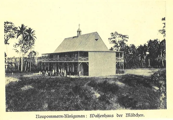 Old photograph of a two-story wooden mission house with a veranda and a small cross on the roof, among palm trees on New Britain