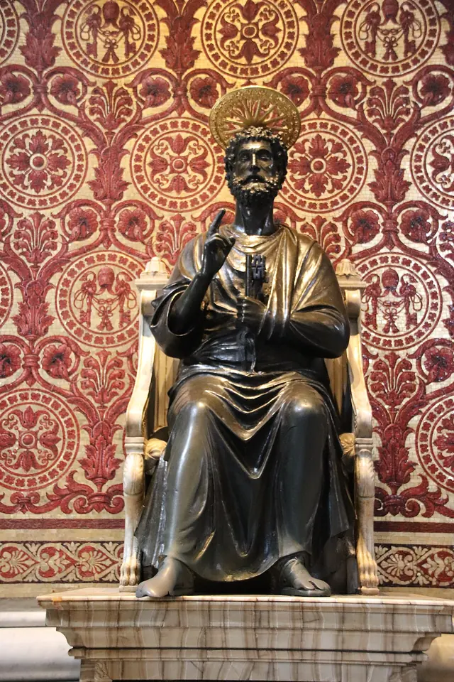 Dark bronze statue of St. Peter seated on a marble chair, raising his right hand in blessing and holding keys in his left, against a red patterned mosaic background