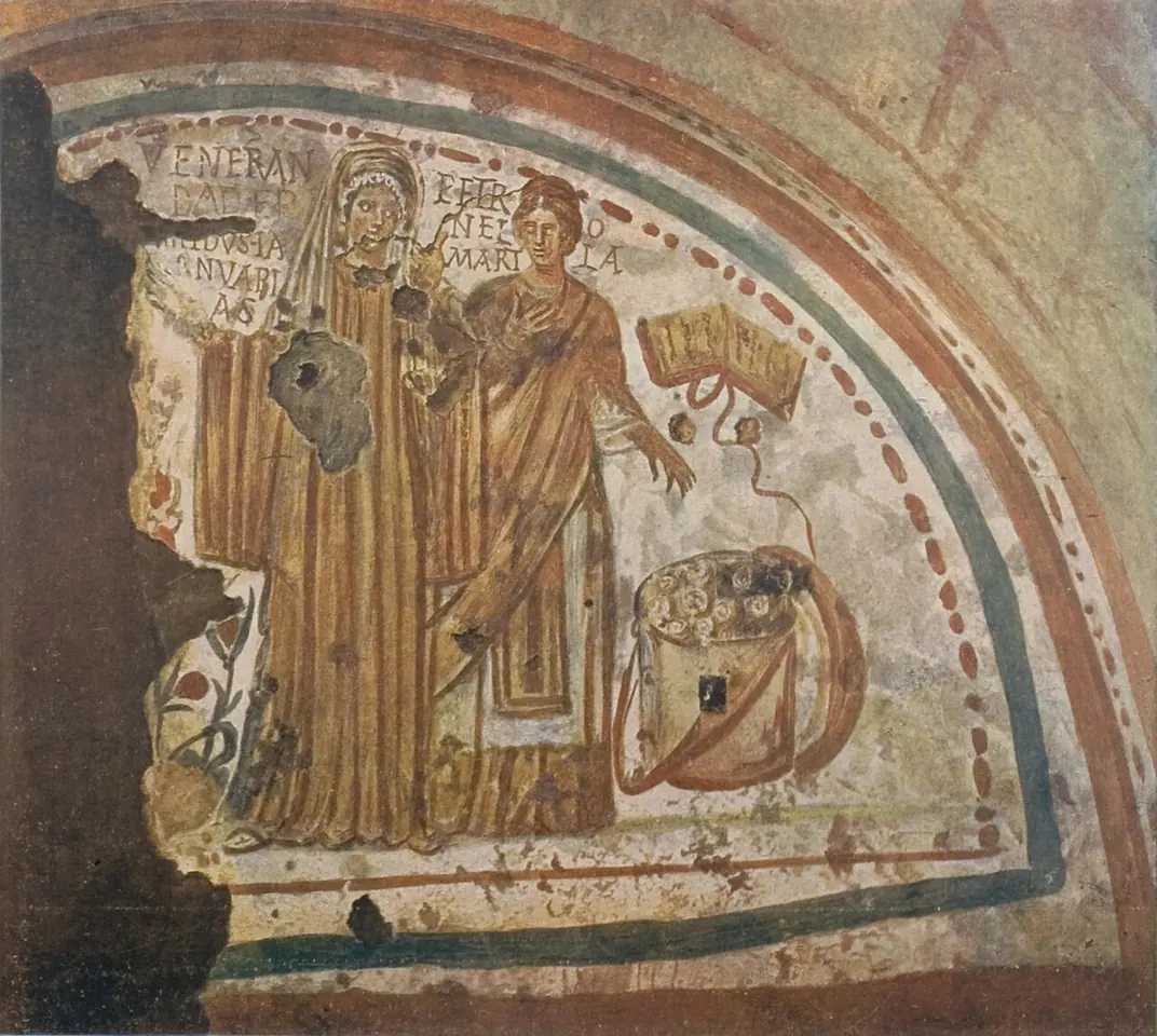 Faded catacomb fresco of two women in long robes, the saint at the right pointing the way, with an open book and a box of scrolls beside them and their names painted above