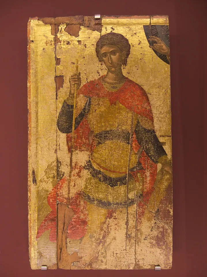 Worn Byzantine icon on gold of a young soldier saint in a red cloak and armor, holding a slender cross-topped staff, with the hand of God blessing from the upper corner