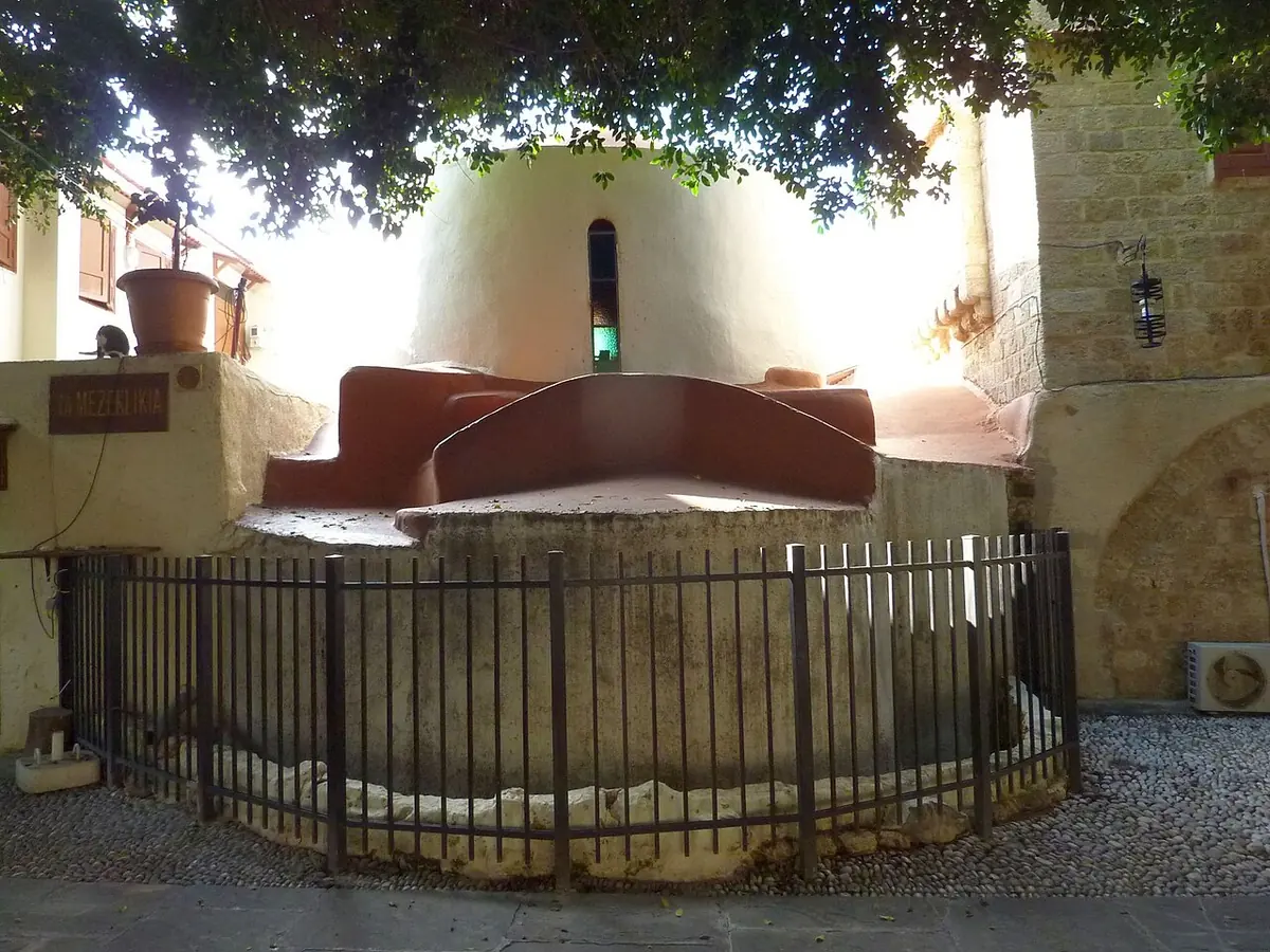 The low, rounded domes and old stone walls of the small Byzantine church of St. Phanourios in the Old Town of Rhodes, behind a black iron railing under a shade tree