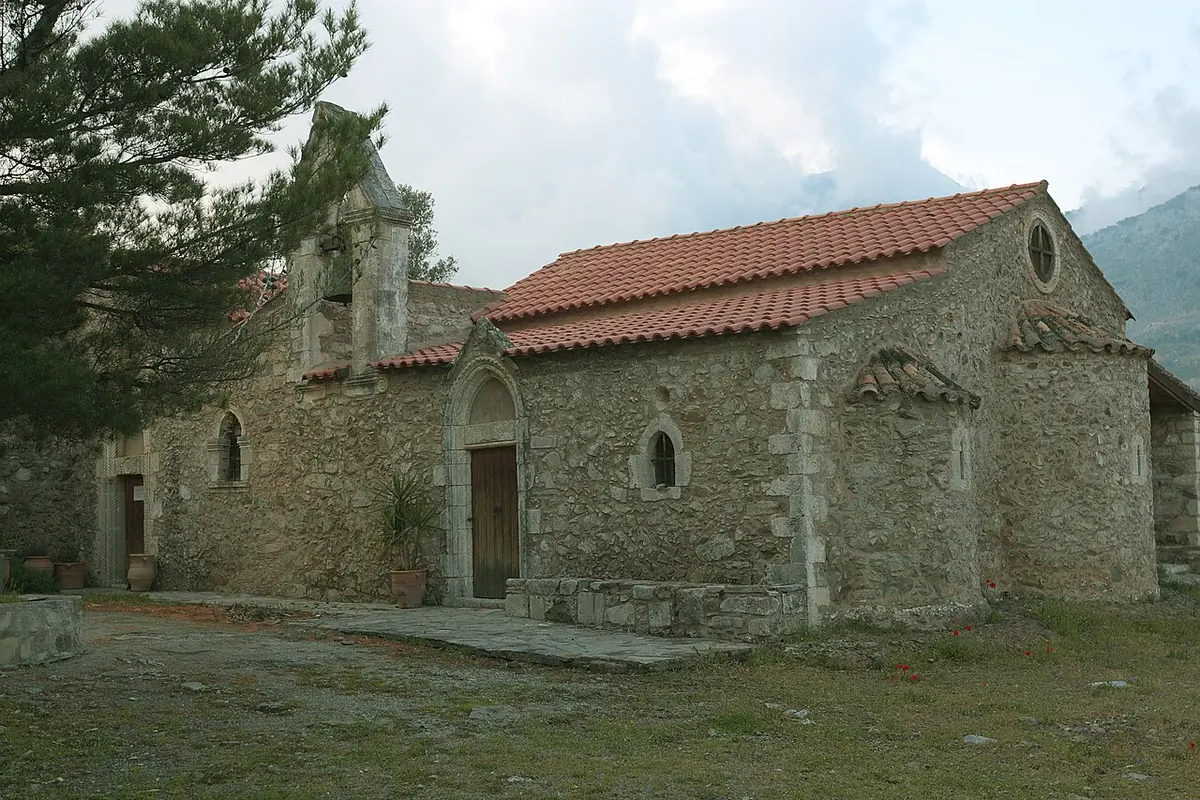 A long stone church with a red tile roof and arched doorway on a grassy slope with mountains behind, the old monastery church of Valsamonero in Crete