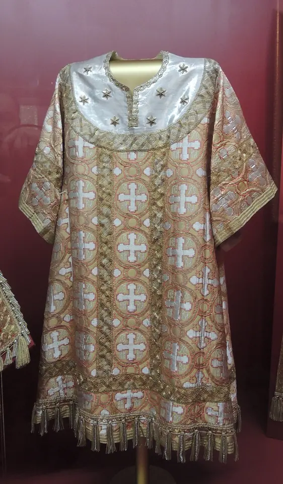 A pale gold brocade bishop's vestment woven with crosses, with a white and gold collar, displayed on a stand in a museum case