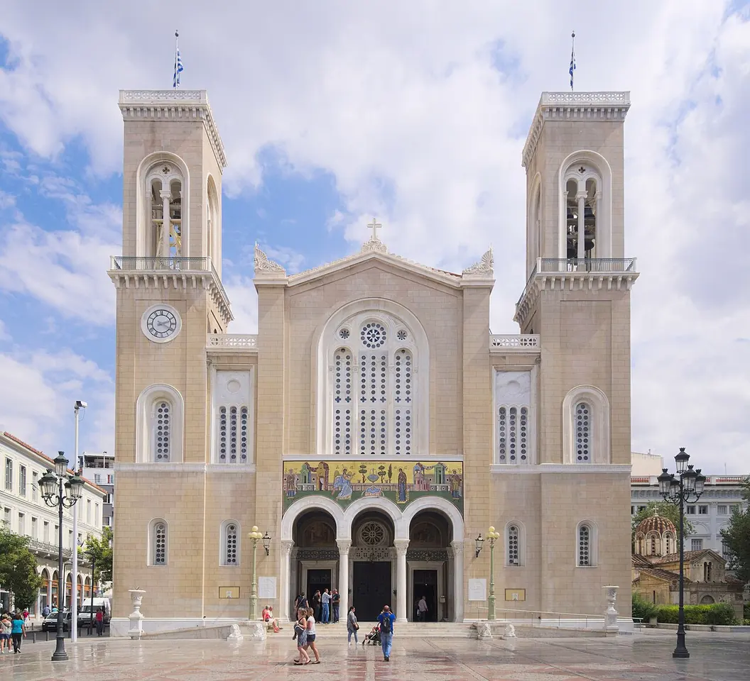 Pale stone cathedral front with two tall bell towers, a rose window and a gold mosaic over the arched entrance, under a cloudy sky