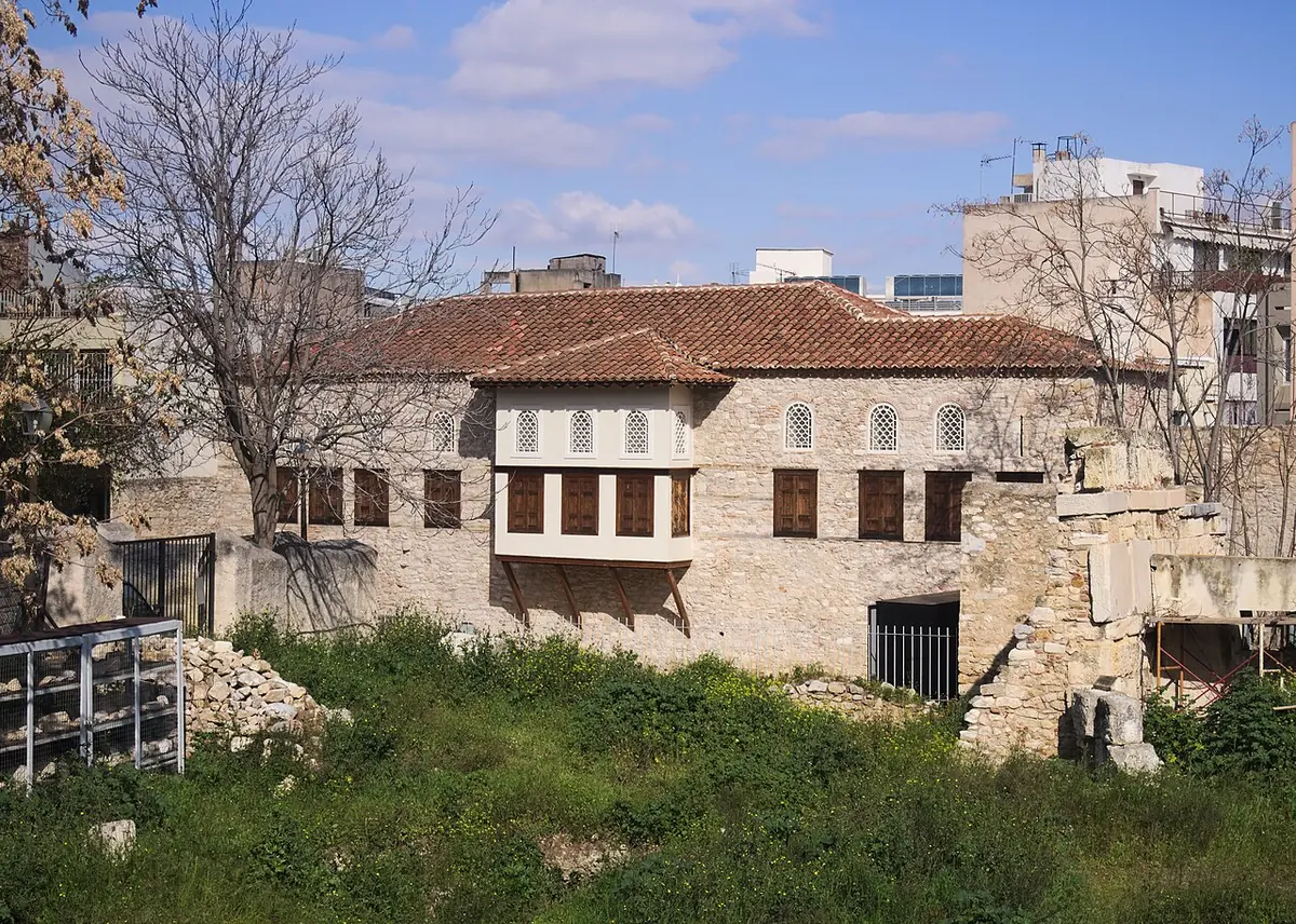 Long two-story stone house with a red-tiled roof and a wooden enclosed balcony, seen across a grassy lot in central Athens