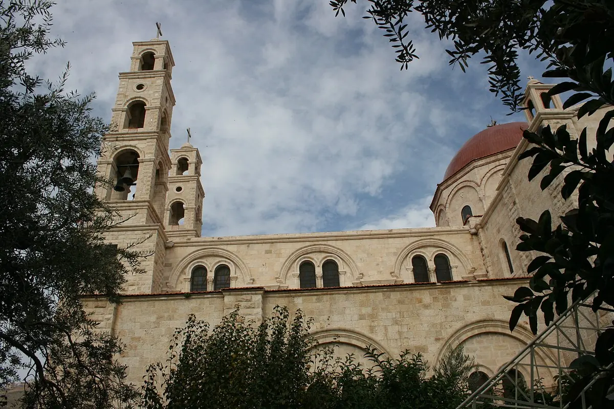 Pale stone church with a tall bell tower and a red dome seen through trees against a cloudy sky