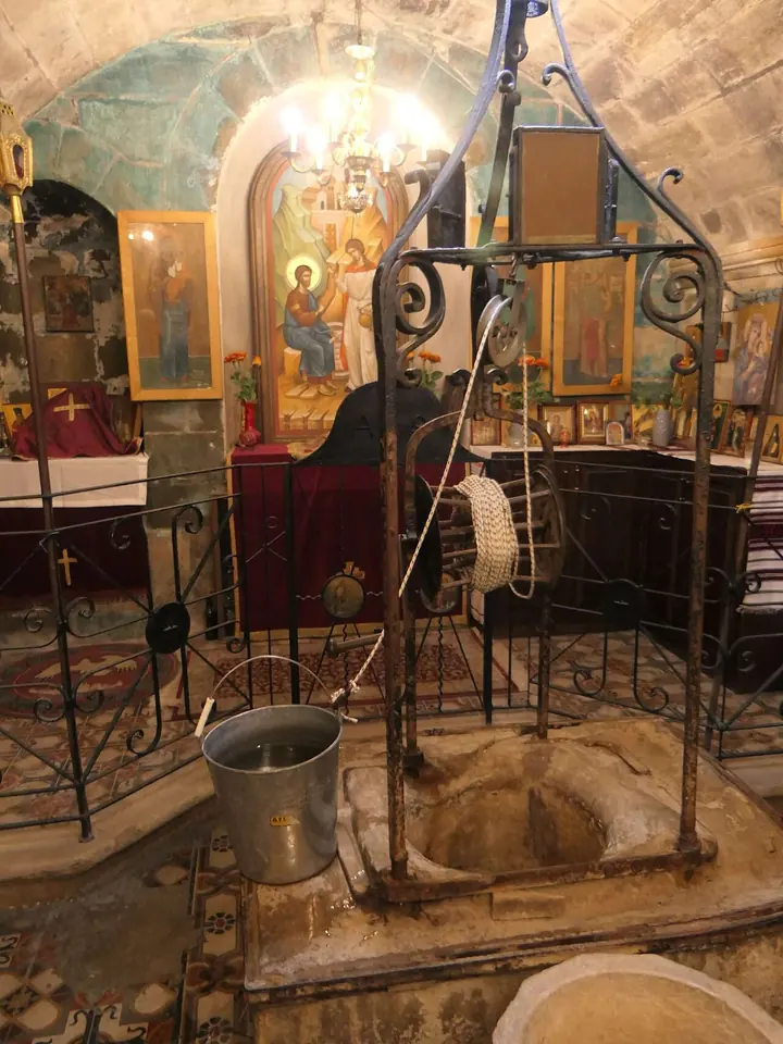 A stone wellhead with a metal frame and pulley and a bucket, in a crypt with icons, lamps and an altar behind