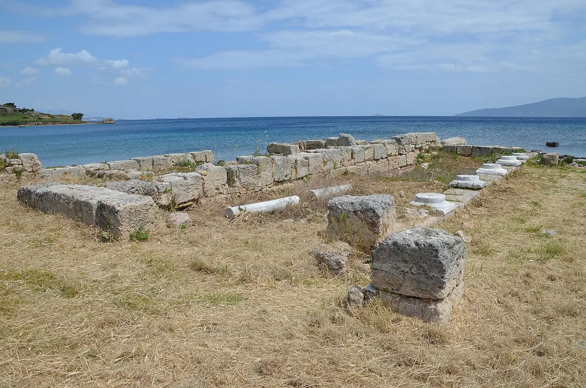 Low stone walls and a paved central aisle of a ruined early Christian basilica on a grassy shore beside the blue water of the Saronic Gulf