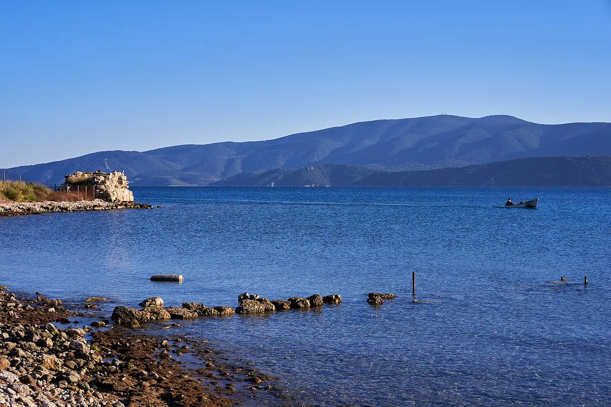 Calm blue bay with the stone foundations of the ancient harbor at the waterline and the ruin of a square tower on a point, under clear sky, with mountains across the water