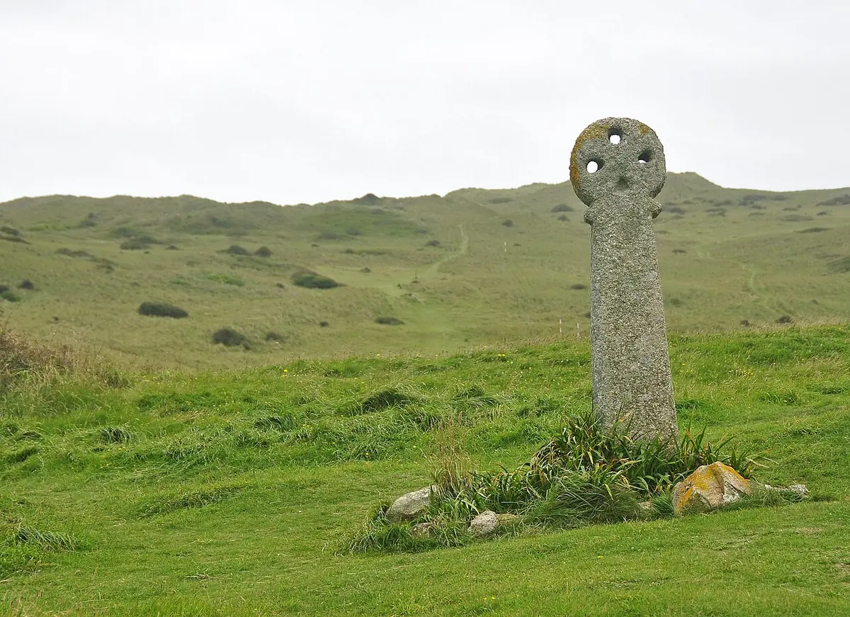 A tall weathered granite cross with a round wheel head standing alone on green, grassy dunes